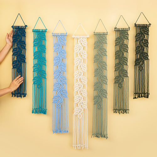 Six macrame wall hangings in different colors are displayed against a warm beige background. A person's hands are visible on the left, holding the dark blue one to show scale.