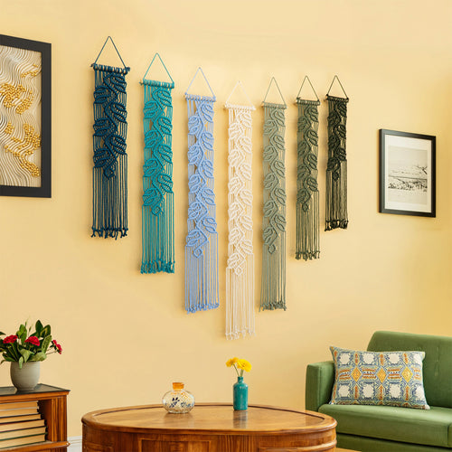 Six woven macrame wall hangings in various colors (blue, teal, light blue, white, sage green, and dark green) are displayed on a beige wall in a living room setting.