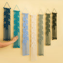 Six woven macrame wall hangings in different colors are displayed on a beige wall, with a person's hand reaching toward the teal one on the left.