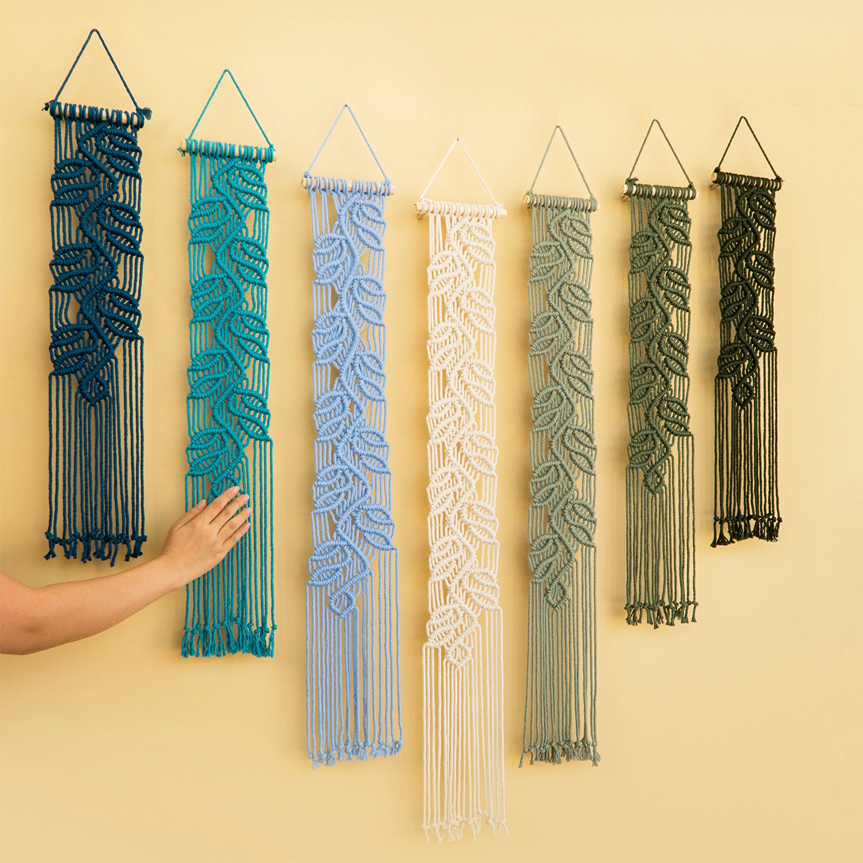 Six woven macrame wall hangings in different colors are displayed on a beige wall, with a person's hand reaching toward the teal one on the left.