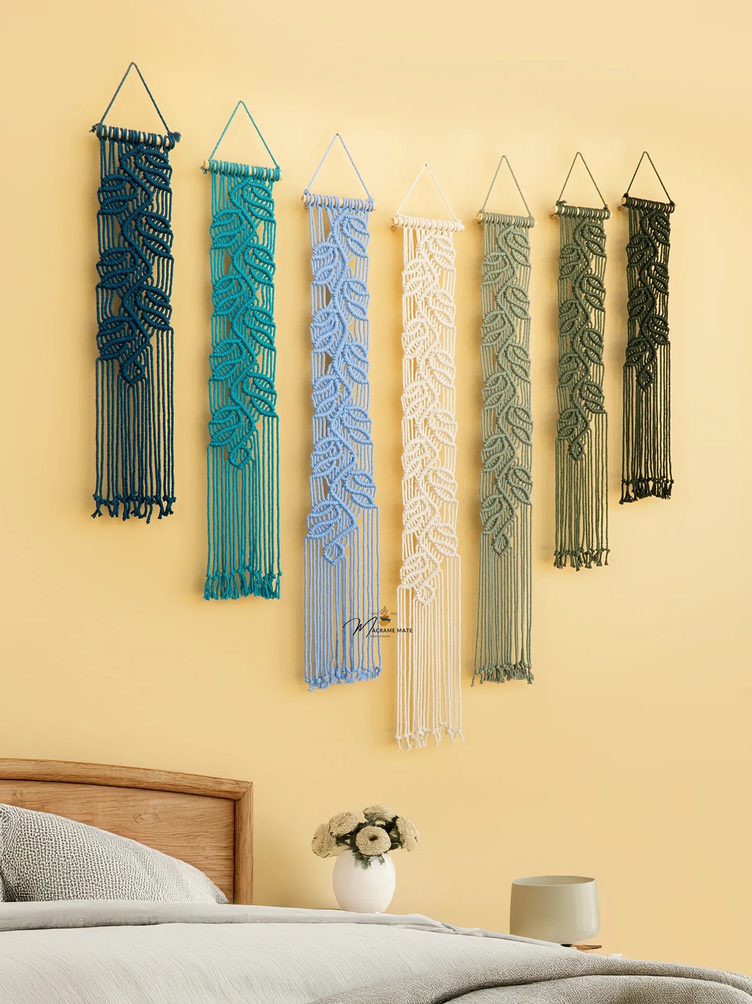 Five macrame wall hangings in various colors on a yellow wall above a bed.