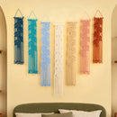 Six woven macrame wall hangings in blue, teal, light blue, cream, beige, pink, and terracotta are displayed on a warm beige wall above a green headboard in a styled bedroom setting.