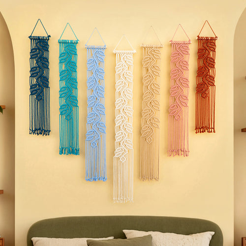 Six woven macrame wall hangings in blue, teal, light blue, cream, beige, pink, and terracotta are displayed on a warm beige wall above a green headboard in a styled bedroom setting.