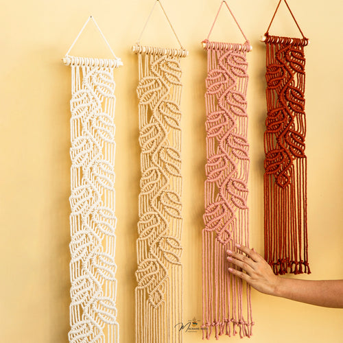 Four macrame wall hangings in white, cream, dusty pink, and terracotta red, displayed vertically against a warm beige wall. A hand is shown touching the pink macrame.