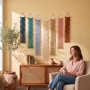 A woman sits in a beige armchair holding a mug in a warm, sunlit room, with six colorful macrame wall hangings displayed on a yellow wall above a wooden cabinet.