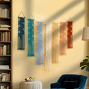 Six macrame wall hangings in blue, teal, light blue, cream, peach, and terracotta are displayed in a row on a beige wall in a styled living room setting with a bookshelf, blue armchair, and plant.