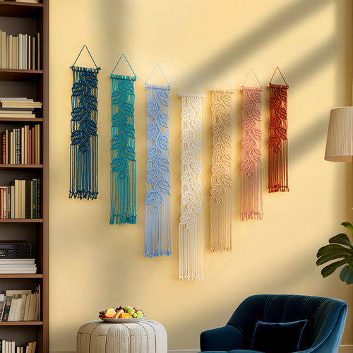 Six macrame wall hangings in blue, teal, light blue, cream, peach, and terracotta are displayed in a row on a beige wall in a styled living room setting with a bookshelf, blue armchair, and plant.