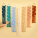 Six macrame wall hangings in different colors are displayed against a warm beige wall. A person's hand is visible on the left, touching the dark blue hanging.