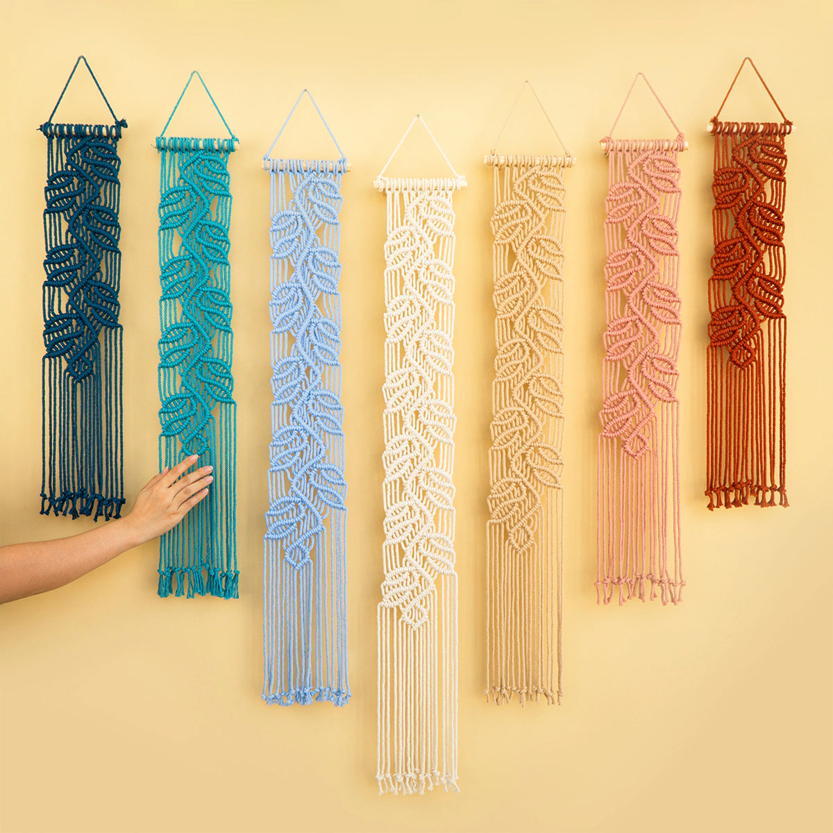 Six macrame wall hangings in different colors are displayed against a warm beige wall. A person's hand is visible on the left, touching the dark blue hanging.