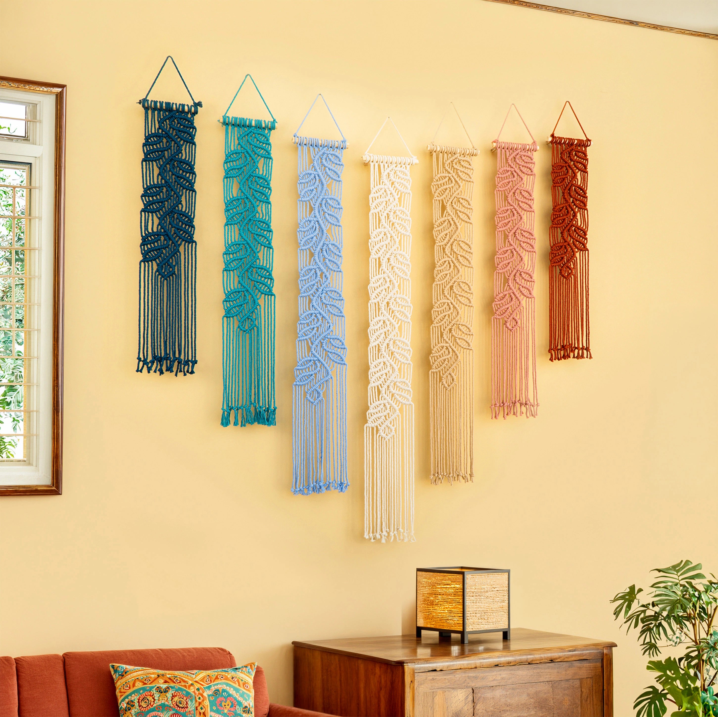 Six macrame wall hangings in different colors are displayed on a beige wall in a styled room setting with a wooden cabinet, plant, and orange sofa.