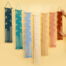 Six macrame wall hangings in different colors are displayed against a solid pale yellow background. A person's hand is visible on the right, holding the red-orange one.