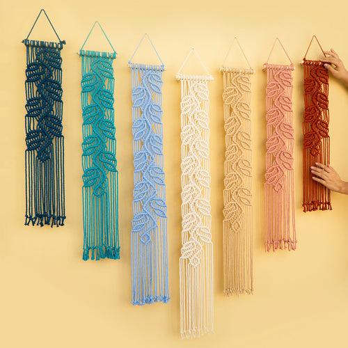 Six macrame wall hangings in different colors are displayed against a solid pale yellow background. A person's hand is visible on the right, holding the red-orange one.