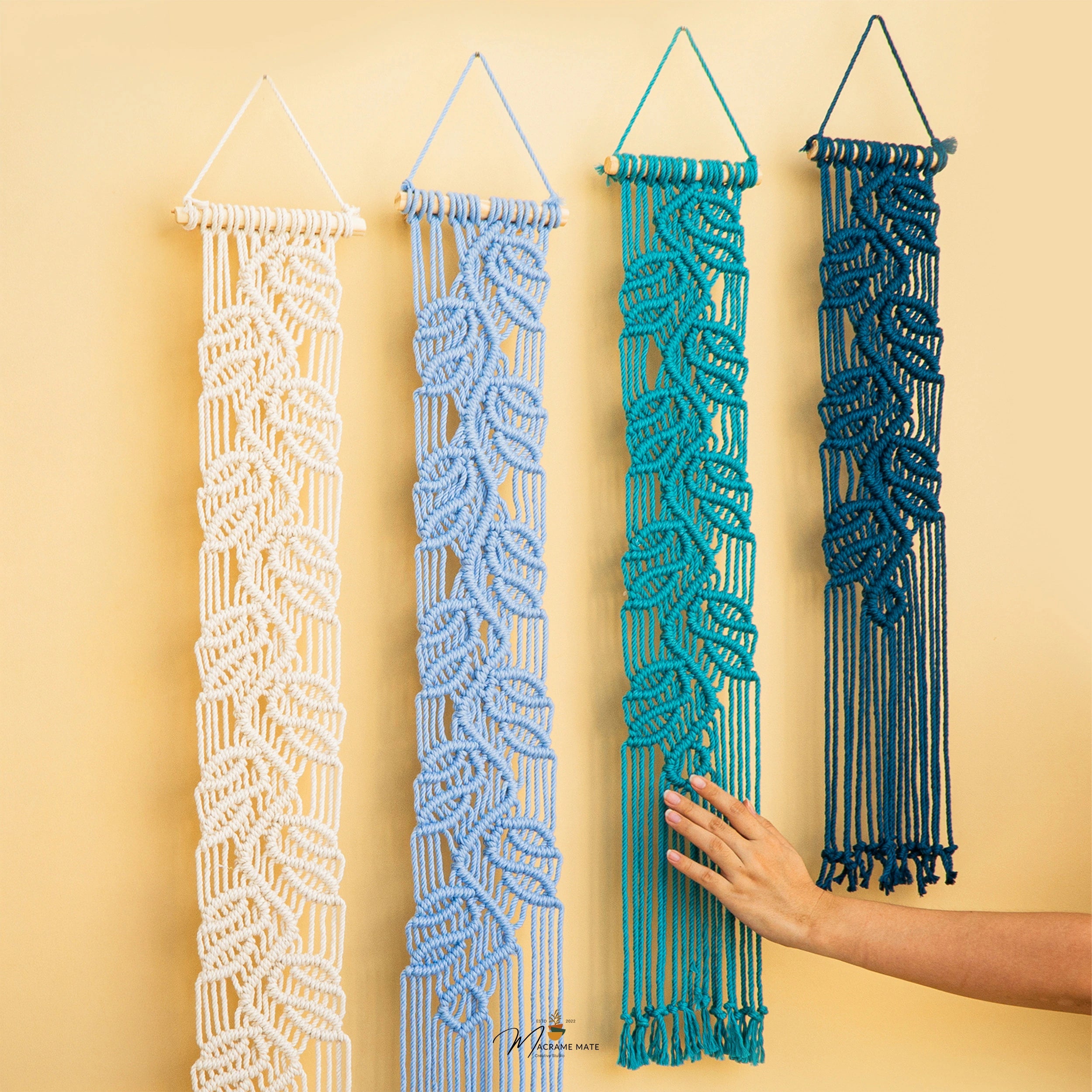 Four macrame wall hangings in white, light blue, teal, and dark blue are displayed against a warm beige wall. A hand is touching the teal hanging to show its texture and scale.