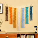 Six leaf-patterned macrame wall hangings in black, orange, mustard, cream, light blue, teal, and navy, displayed in a row on a beige wall above a wooden bookshelf.
