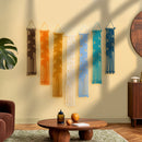 Six woven macrame wall hangings in brown, orange, cream, light blue, teal, and navy are displayed on a beige wall in a styled living room setting with a wooden coffee table, plant, and warm ambient lighting.