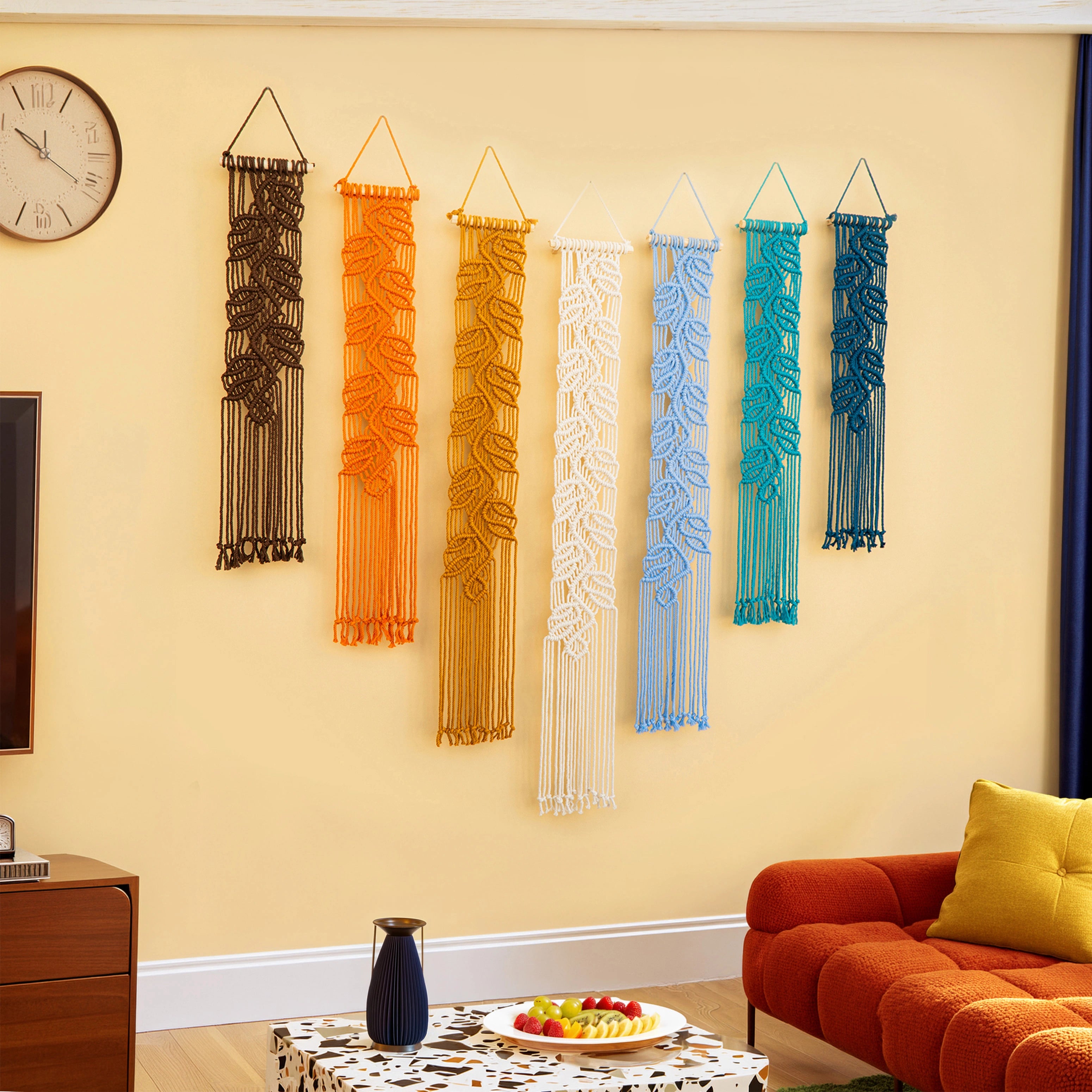 Six macrame wall hangings in black, orange, yellow, white, light blue, and teal are displayed in a row on a beige wall in a living room setting with a red sofa, a clock, and a fruit stand.