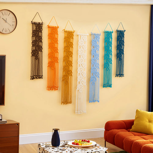 Six macrame wall hangings in black, orange, yellow, white, light blue, and teal are displayed in a row on a beige wall in a living room setting with a red sofa, a clock, and a fruit stand.