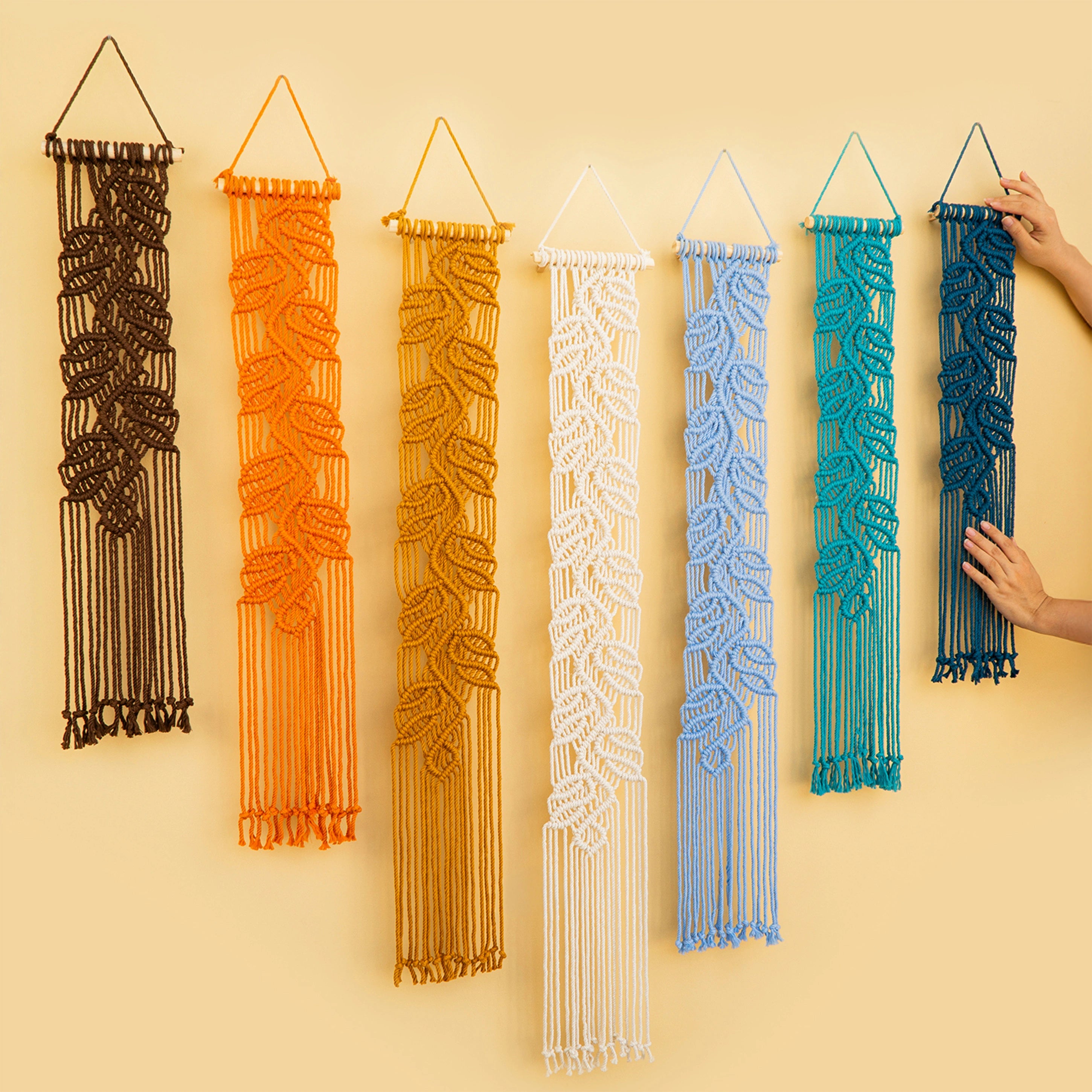 Six macrame wall hangings in dark brown, orange, mustard yellow, white, light blue, teal, and navy blue are displayed on a warm beige wall. Two hands are visible holding the edges of the teal and navy blue hangings, demonstrating scale and wearability.