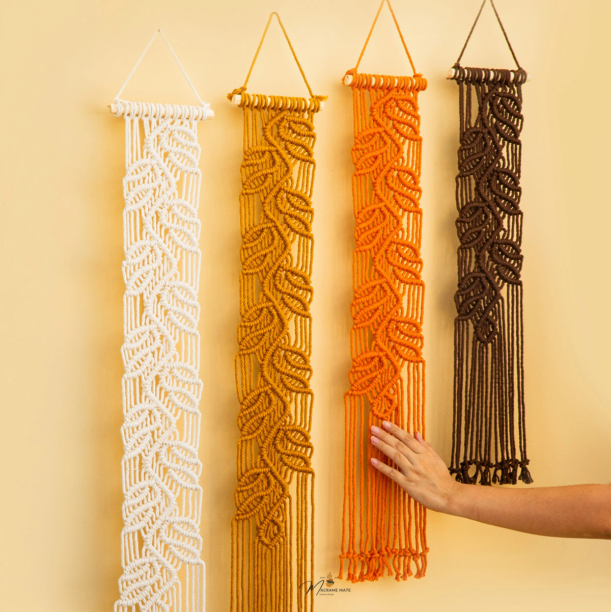 Four vertical macrame wall hangings in white, mustard yellow, bright orange, and dark brown, displayed against a warm beige wall. A person's hand is touching the orange hanging, and a small logo watermark appears at the bottom center.