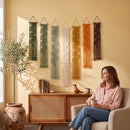 A woman sits in a light beige armchair holding a mug in a warm, sunlit room. Above her, seven boho-style macrame wall hangings in green, white, yellow, and brown are displayed on a beige wall above a wooden credenza.