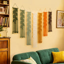 Six macrame wall hangings in green, blue, white, orange, and brown are displayed on a beige wall above a green sofa in a cozy living room setting.