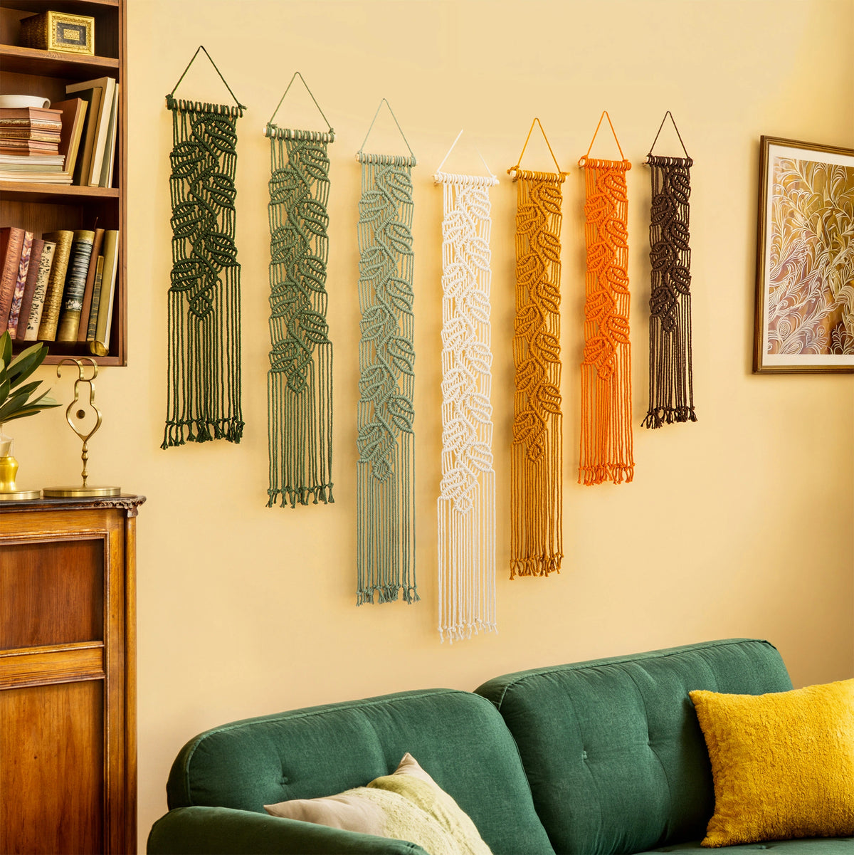 Six macrame wall hangings in green, blue, white, orange, and brown are displayed on a beige wall above a green sofa in a cozy living room setting.