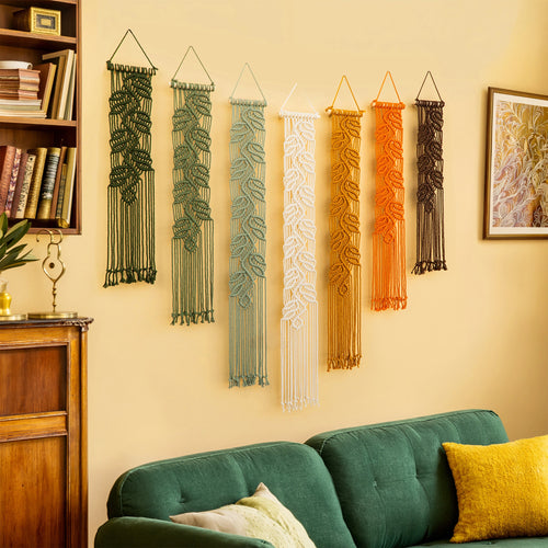 Six macrame wall hangings in green, blue, white, orange, and brown are displayed on a beige wall above a green sofa in a cozy living room setting.