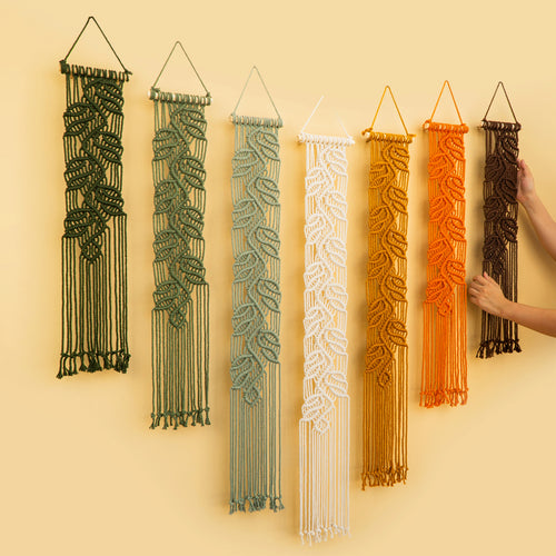 Six macrame wall hangings with leaf patterns in dark green, sage green, white, mustard yellow, orange, and brown, displayed on a warm beige wall. A person's hand is visible on the right adjusting one of the orange hangings.