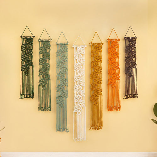 Set of six macrame wall hangings in various colors on a yellow wall.