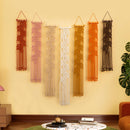 Six macrame wall hangings in different colors are displayed on a yellow wall in a styled living room setting with a green rug, coffee table, and orange sofa.