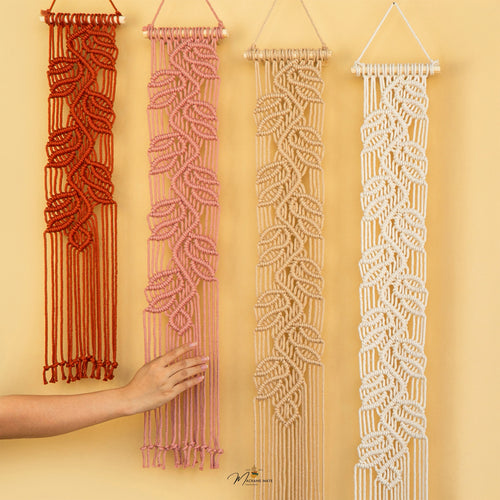 Four macrame wall hangings in rust red, dusty pink, beige, and off-white, displayed against a warm yellow wall. A hand is shown touching the pink hanging, and a small 'Mikayla's' logo is visible at the bottom.