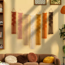 Six macrame wall hangings in warm neutral and earthy tones are displayed on a beige wall above a brown sofa with textured throw pillows in a styled living room setting.