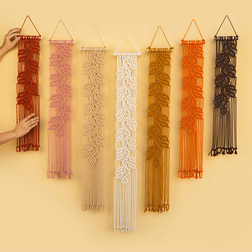 Six macrame wall hangings in warm earthy tones are displayed against a solid pale yellow background. A person's hands are visible on the left, holding the rust-colored hanging.