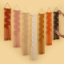 Six macrame wall hangings in different colors are displayed on a warm beige wall. A pair of hands is visible on the right, holding the black one in place.