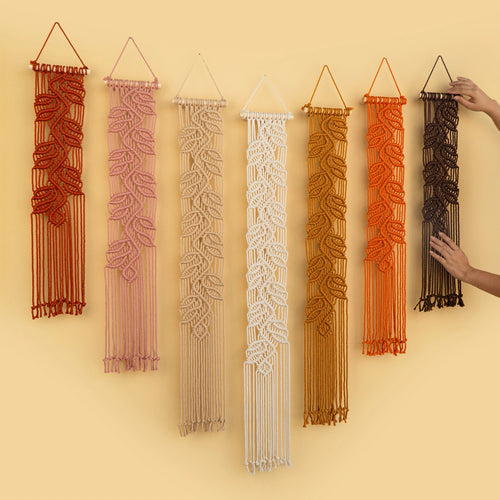 Six macrame wall hangings in different colors are displayed on a warm beige wall. A pair of hands is visible on the right, holding the black one in place.
