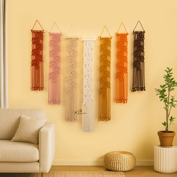 Set of six macrame wall hangings in various colors on a beige wall with a sofa and plant in the foreground.