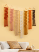 Set of six macrame wall hangings in various colors on a yellow wall above a white sofa.