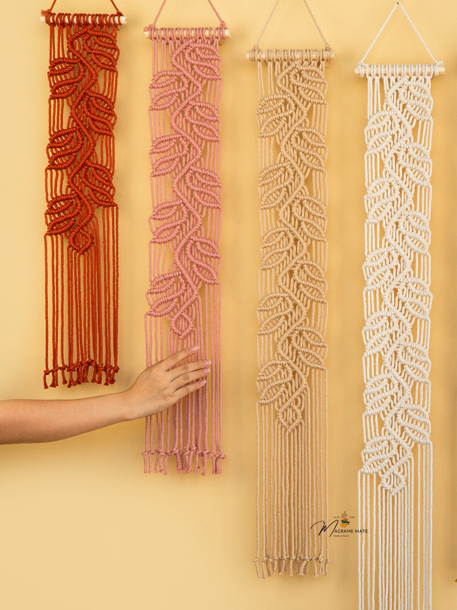 Four macrame wall hangings in different colors on a yellow wall.