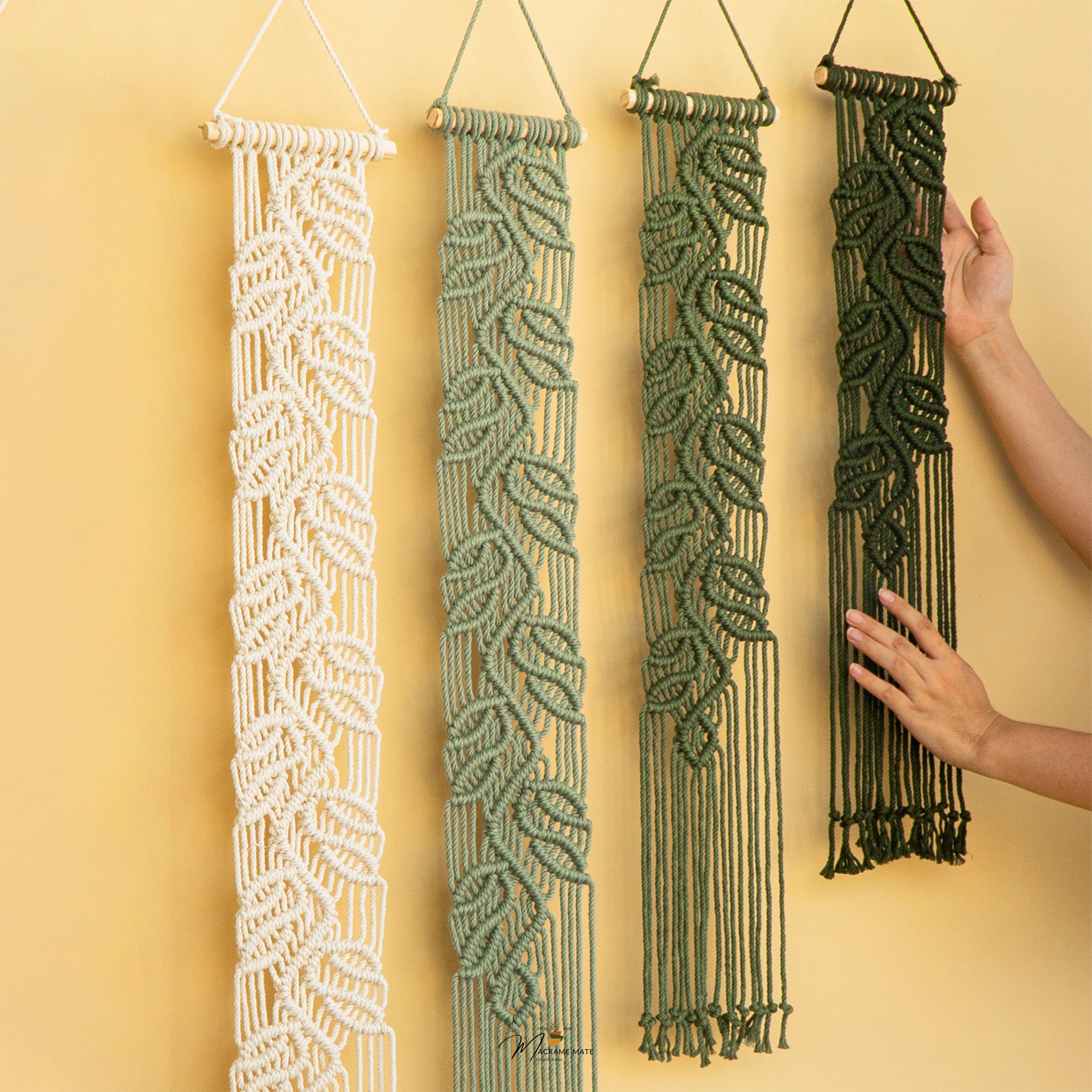 Four leaf-patterned macrame wall hangings in cream, sage green, olive green, and dark forest green, displayed against a warm yellow wall with a person's hands adjusting the rightmost hanging.