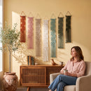 A woman sits in a beige armchair holding a mug in a warm, styled room, with six colorful macrame wall hangings displayed on a yellow wall above a wooden cabinet.
