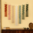 Six woven macrame wall hangings in terracotta, pink, cream, sage green, dusty blue, and forest green, displayed on a beige wall above a dark wooden console table with books and small decor.