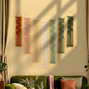 Six macrame wall hangings in terracotta, pink, cream, mint, and forest green are displayed on a beige wall above a green sofa in a styled living room setting.