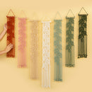 Six leaf-patterned macrame wall hangings in rust red, dusty pink, cream, off-white, sage green, and forest green, displayed against a warm beige background. A hand is visible on the left touching the rust red hanging.