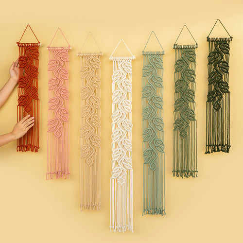 Six leaf-patterned macrame wall hangings in rust red, dusty pink, cream, off-white, sage green, and forest green, displayed against a warm beige background. A hand is visible on the left touching the rust red hanging.