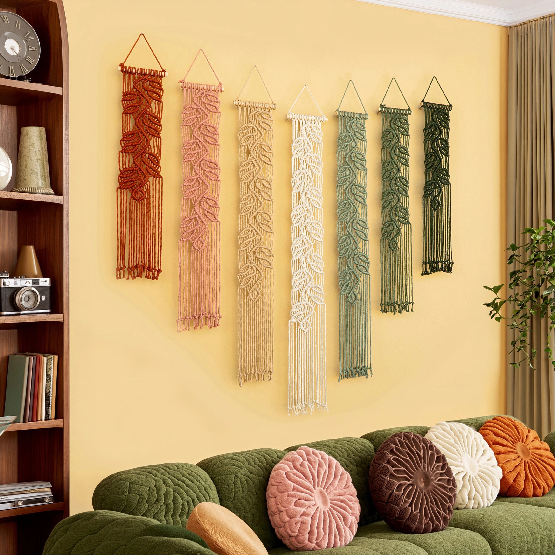 Six macrame wall hangings in earthy tones of terracotta, dusty pink, cream, sage green, mint green, and olive green, displayed on a warm yellow wall above a green sofa with decorative pillows.