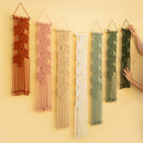 Six macrame wall hangings with leaf patterns in rust, pink, cream, sage green, and forest green, displayed on a warm beige wall. Two hands are visible holding the rightmost hangings to show scale.