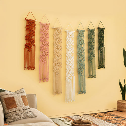 Set of six macrame wall hangings in various colors on a yellow wall.