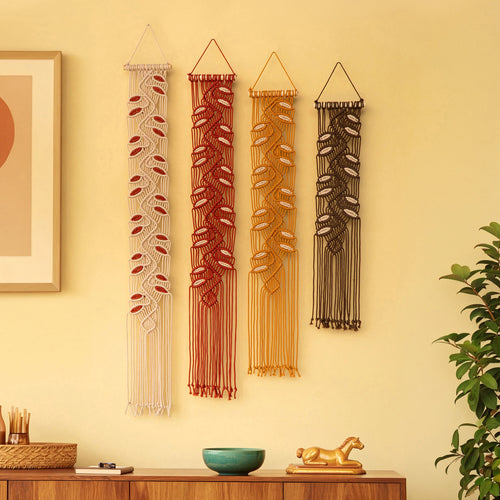 Four bohemian macrame wall hangings with leaf patterns in cream, rust red, mustard yellow, and dark brown, displayed on a warm beige wall above a wooden console table with decorative items.