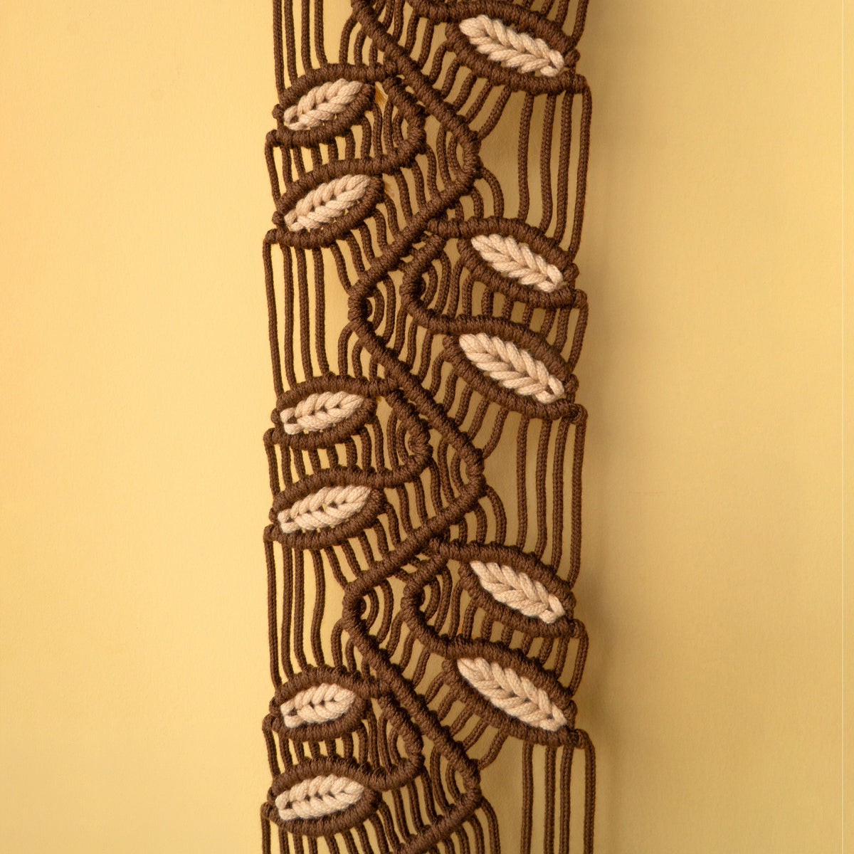 A close-up of a decorative braided cord trim or macrame pattern in dark brown and cream, shown vertically against a solid warm beige background.