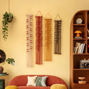 Four bohemian macrame wall hangings in cream, red, mustard, and brown, displayed on a warm yellow wall above a terracotta sofa in a styled living room.